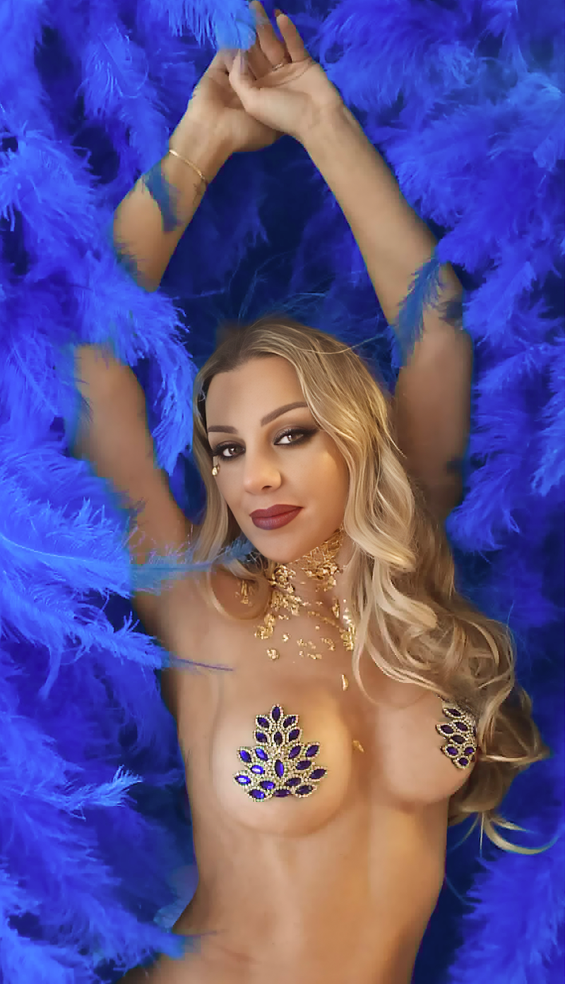 Blonde burlesque performer wearing blue rhinestone pasties poses against vibrant royal blue feathers with raised arms, dramatic makeup, and gold body jewelry in glamorous showgirl-inspired styling.